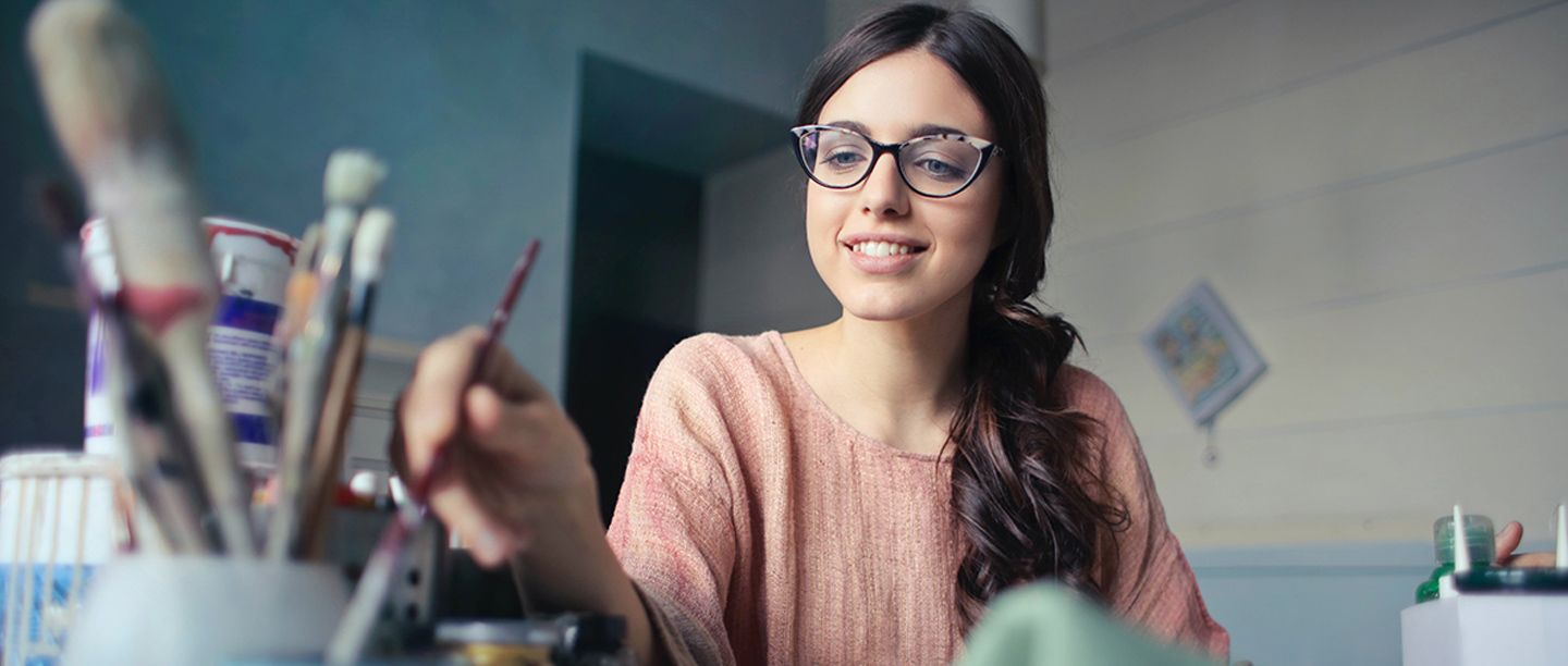 A woman with glasses and long brown hair is smiling while painting. She is holding a paintbrush and wearing a light pink sweater. Various art supplies are visible in the foreground. The room has a calm, muted color scheme.